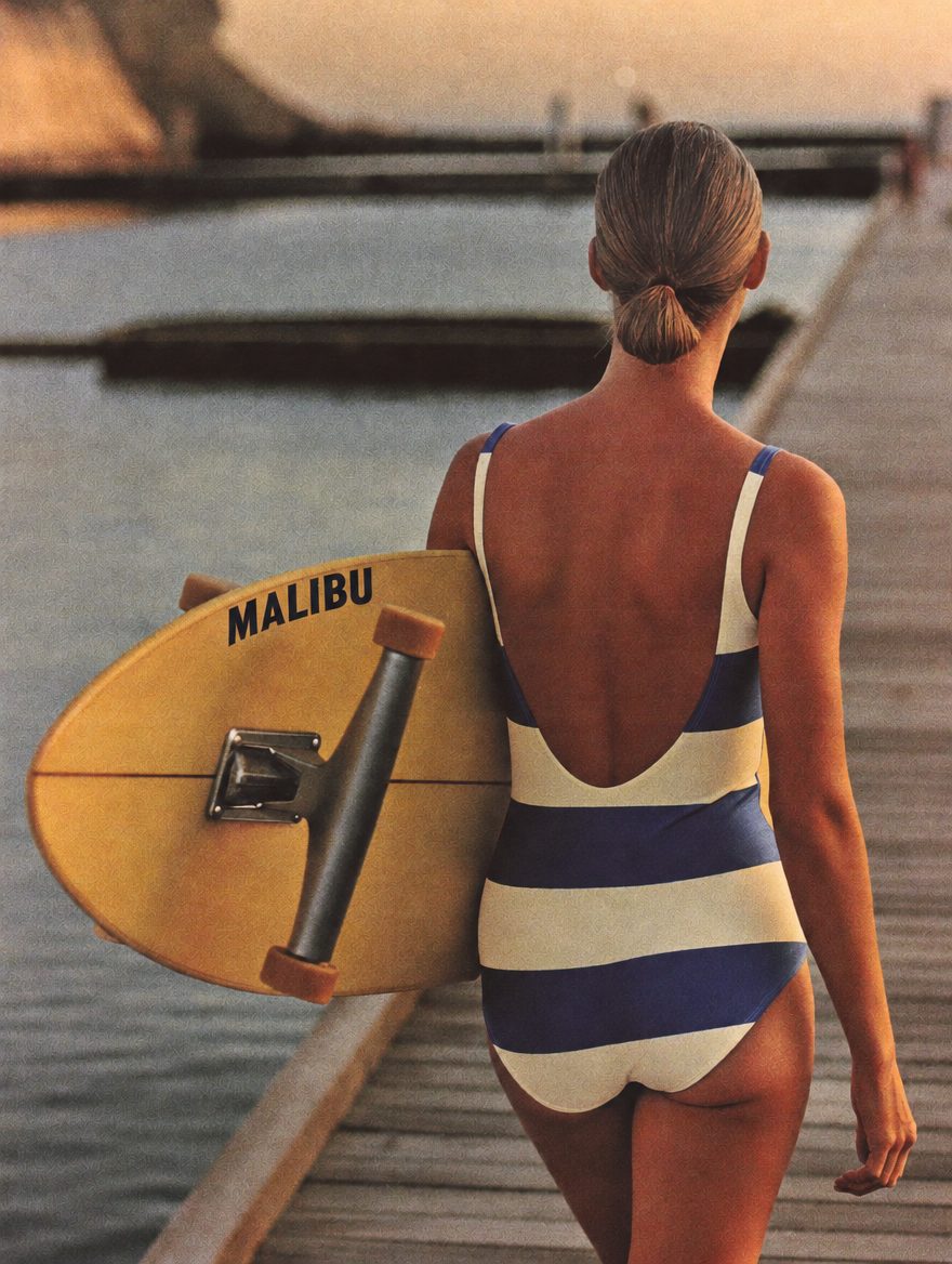 Model in The Malibu striped one-piece carrying a board down the pier
