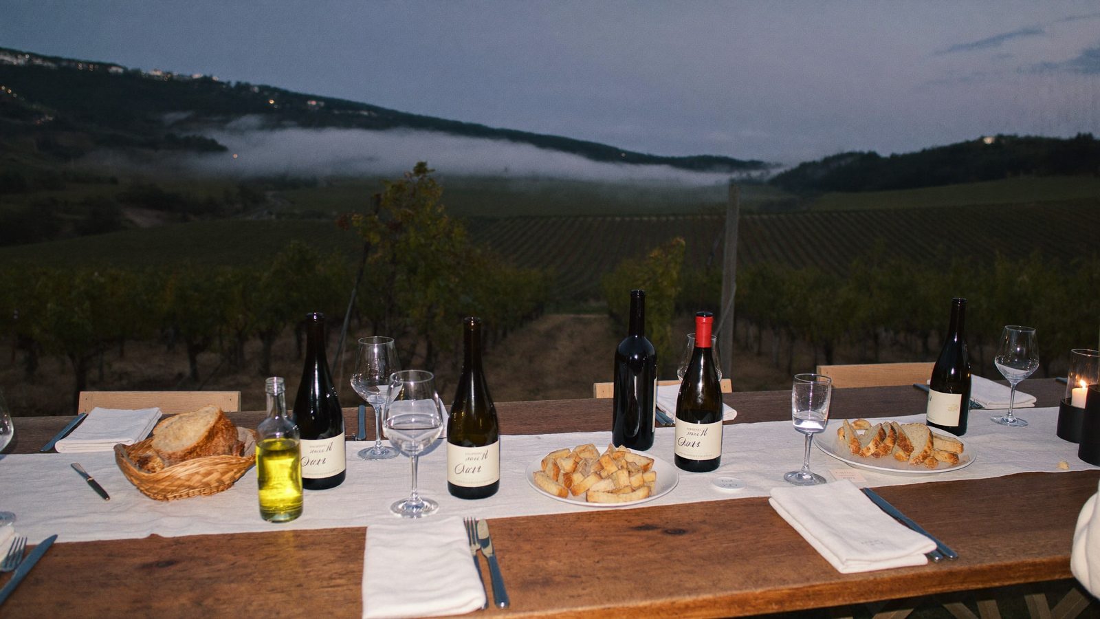 The long table set at the edge of the vines at dusk, fog on the hills