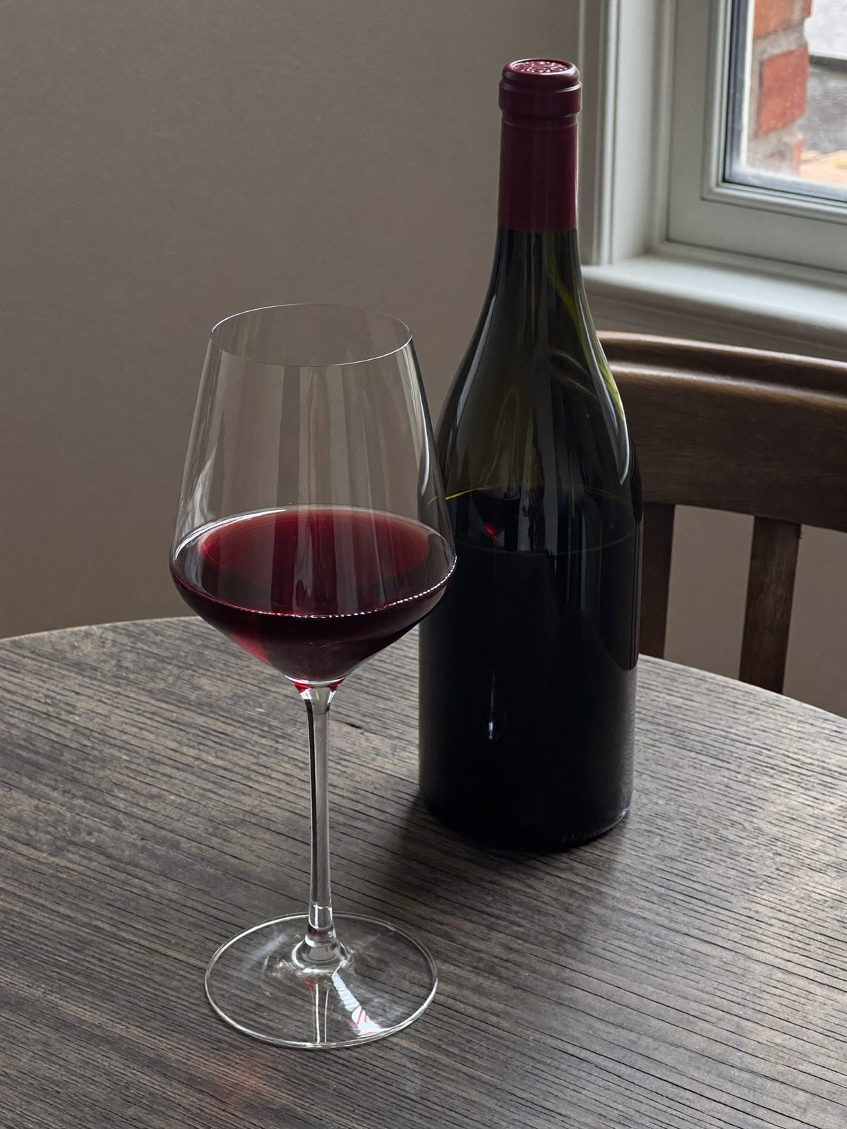 A glass of Pinot Noir beside a dark bottle by a window