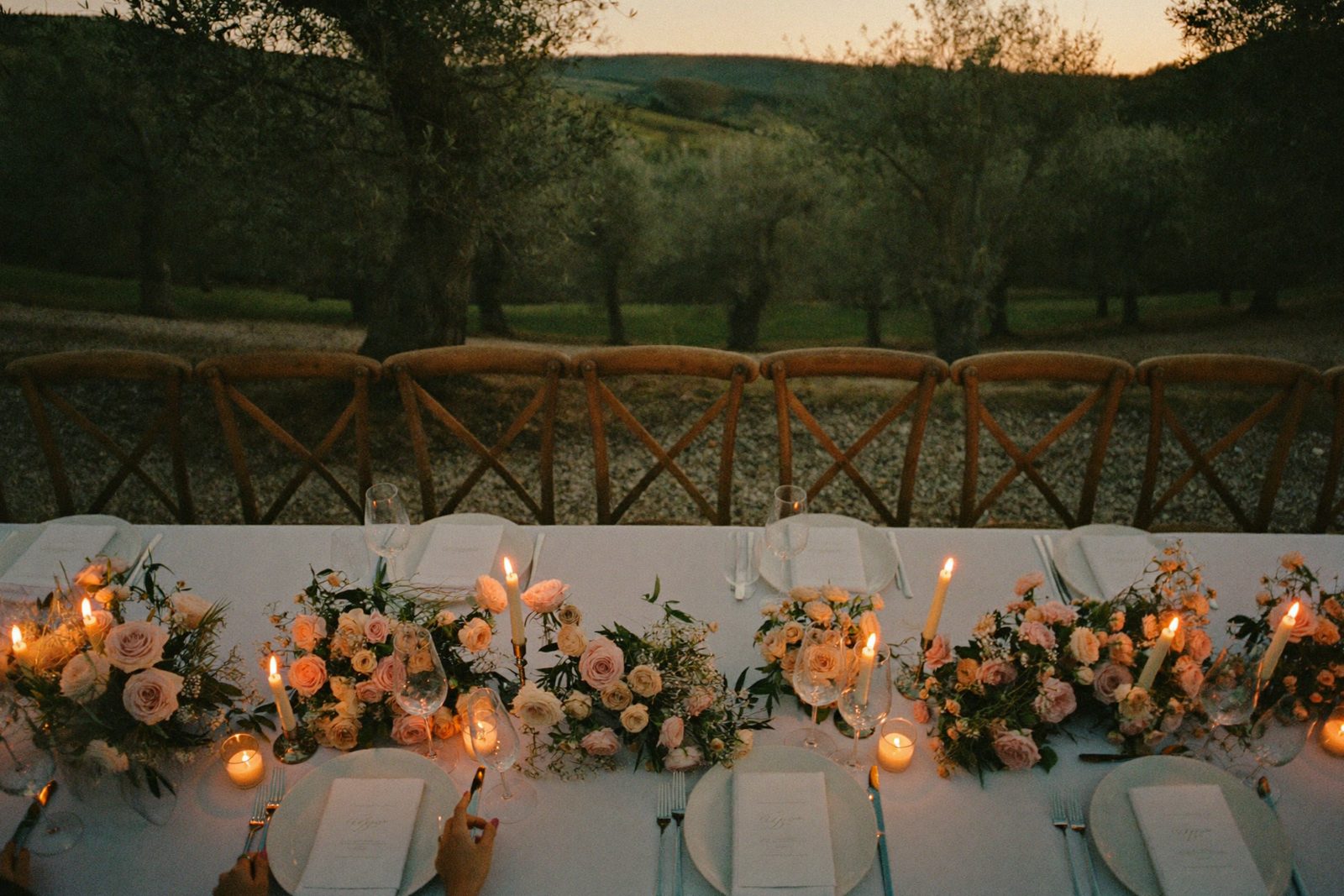 A long candlelit dinner table beneath olive trees at dusk