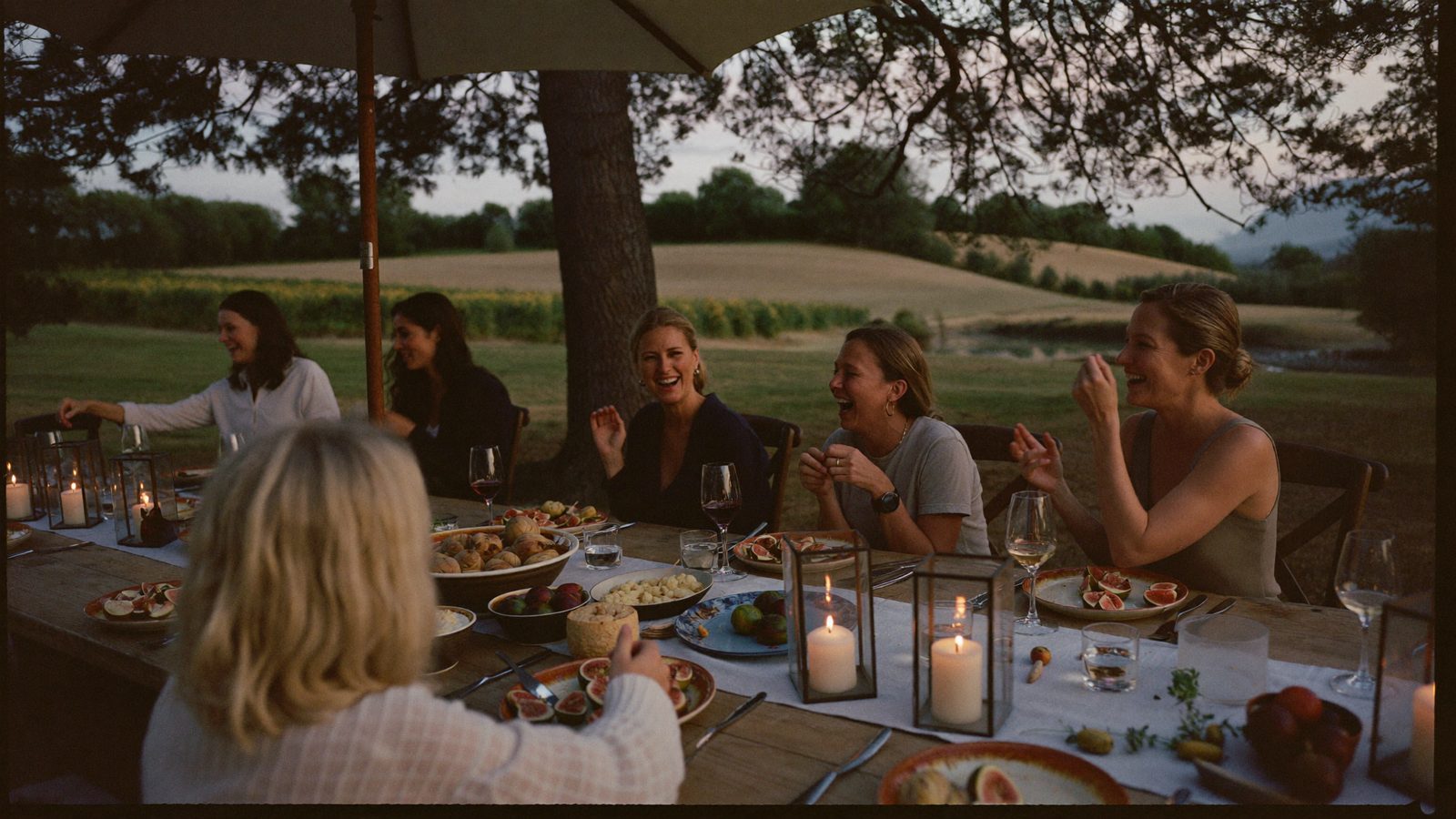 Women laughing at a long candlelit table under the pines at dusk