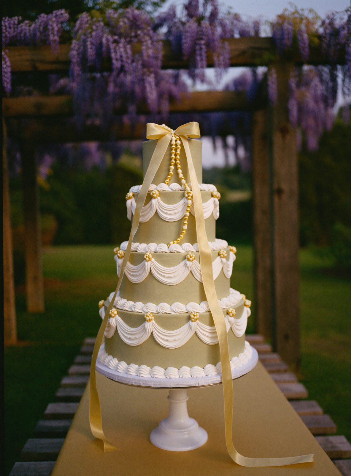 A four tier sage green cake with ivory over piping and sugar sweet peas under a wisteria pergola
