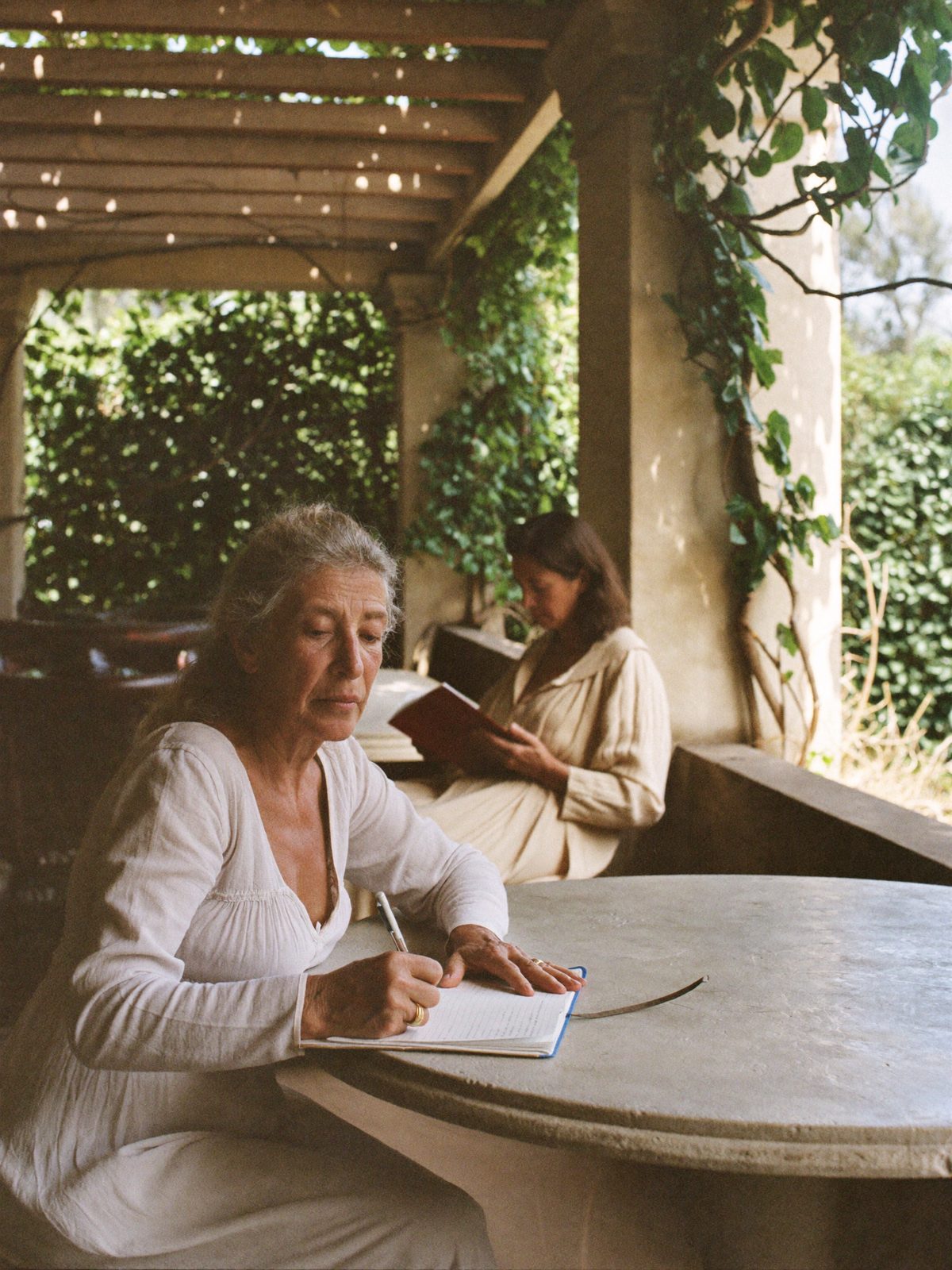 A woman writing at a stone table in a shaded loggia while another reads