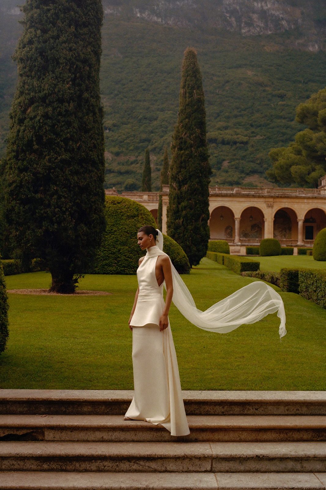 A bride in a sculptural silk column gown, her veil caught in the wind, on the travertine steps of a Roman villa garden